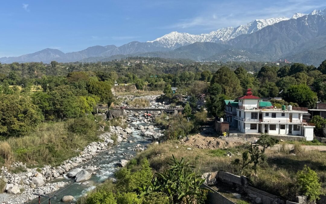 Picturesque landscape of Dharamshala, India showcasing a river, lush greenery, and snowy mountains.
